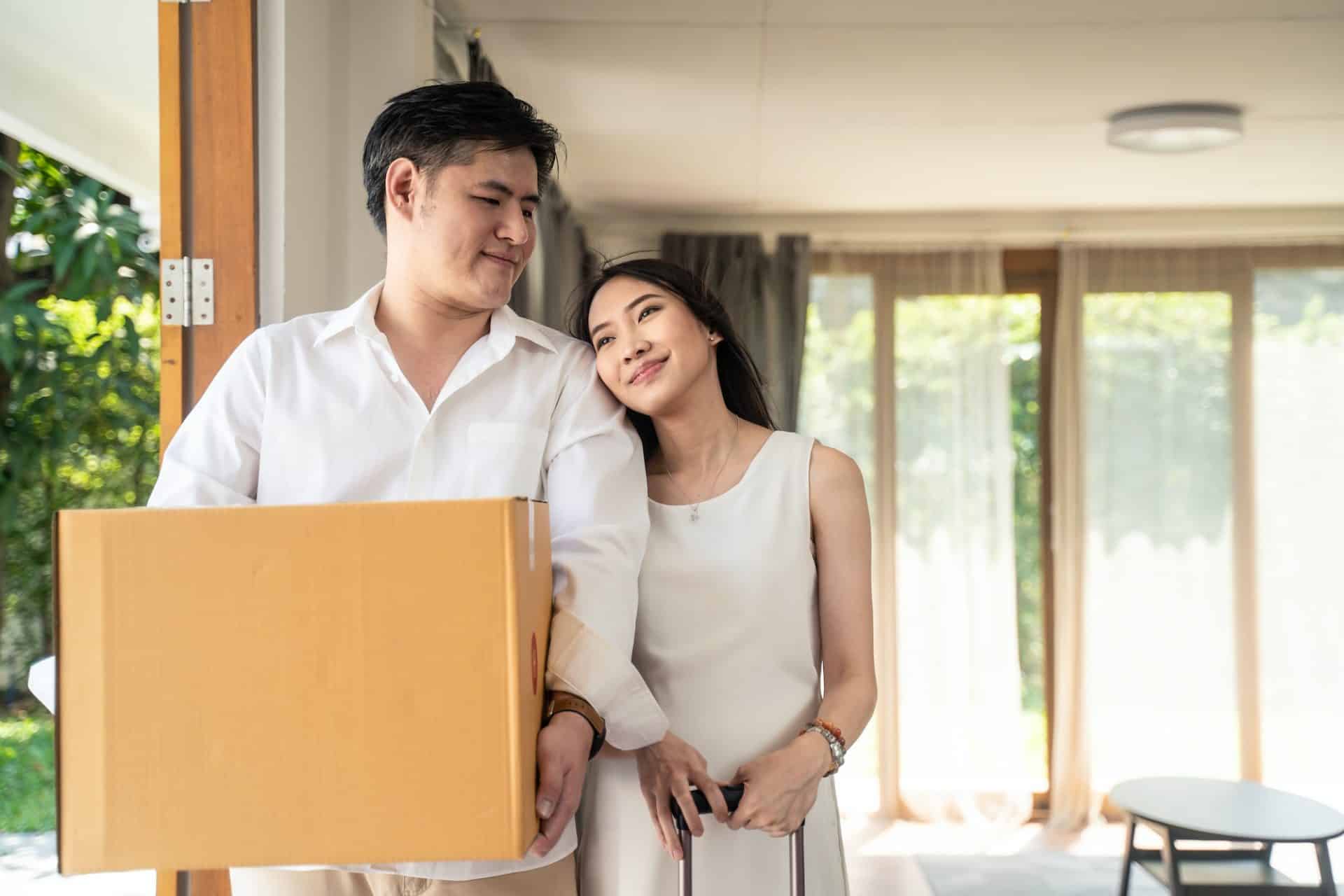 Asian couple moving to new house, man holding big box standing near pregnancy woman